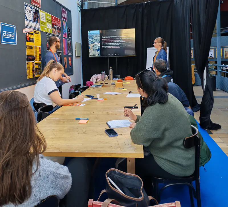 A group of participants sitting at a table, with the facilitators at the front and a slide being shown on a screen.
